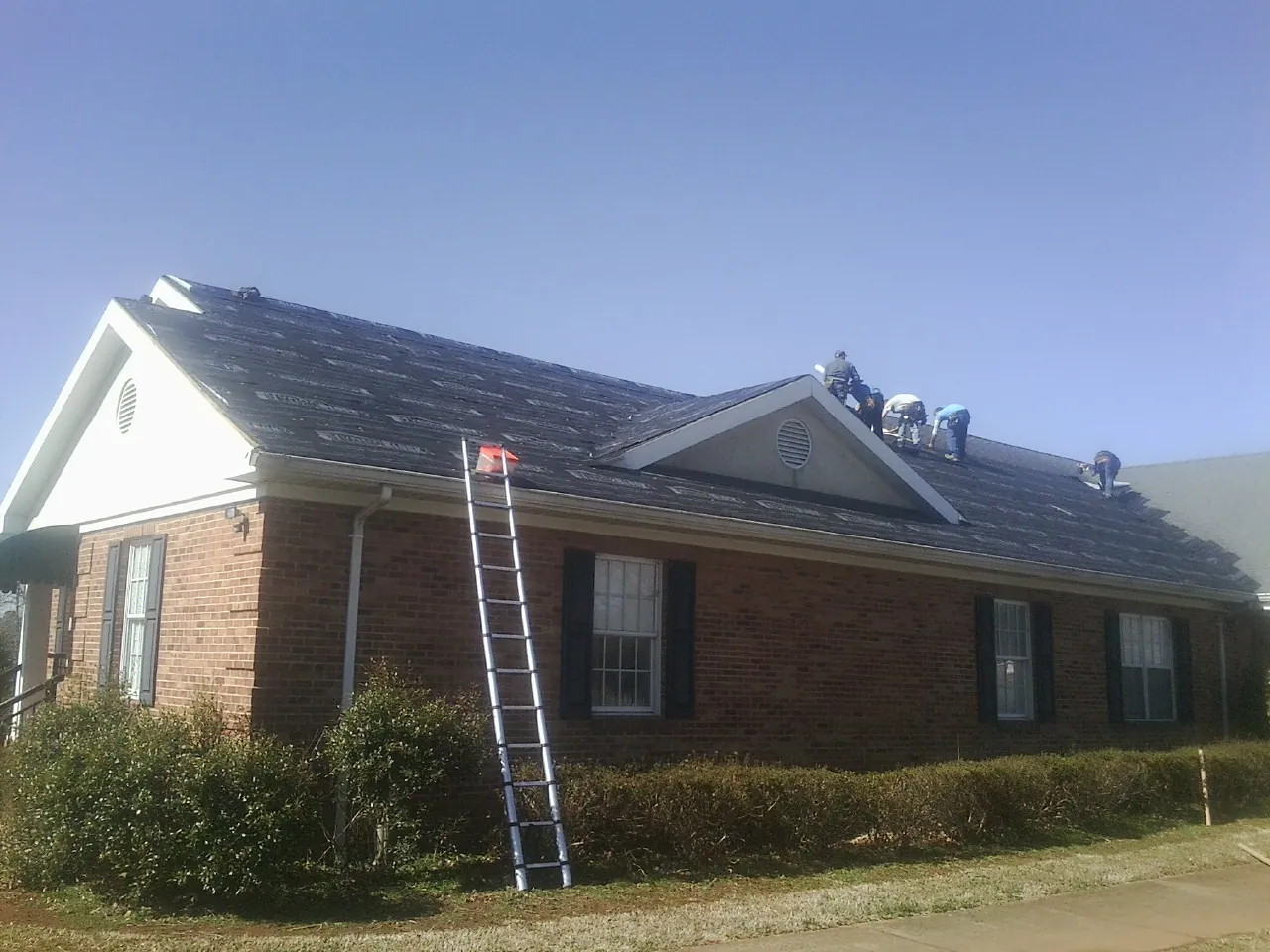 Crew tearing shingles off a brick ranch roof with a ladder set — Metal Roof Repair work by our crew in Buhl