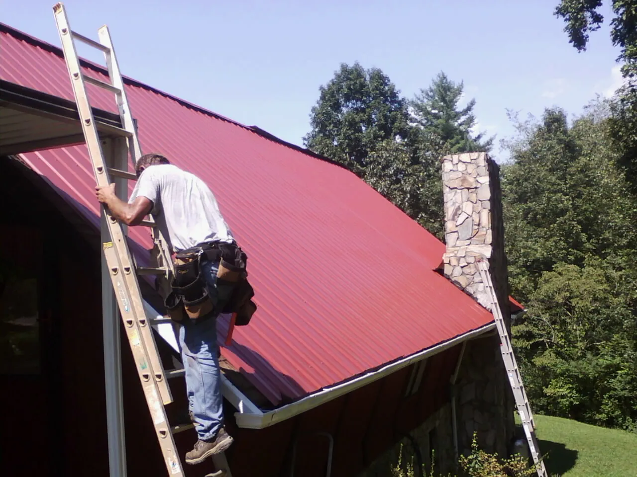 Roofer on a ladder beside a red metal roof and stone chimney — Soffit & Fascia Repair by Roofing Brothers in Buhl
